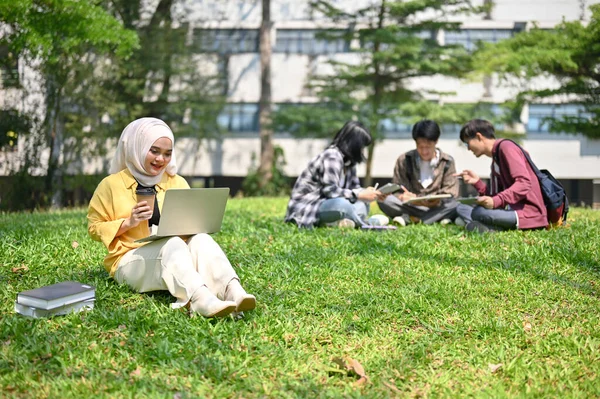 穿着头巾坐在草地上 一边喝着咖啡 一边在校园公园里使用笔记本电脑的年轻而快乐的年轻的亚洲穆斯林女大学生 — 图库照片