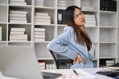 Ofis sendromundan muzdarip yorgun ve kötü Asyalı iş kadını masasında çalışırken sırt ağrısı çekiyor..