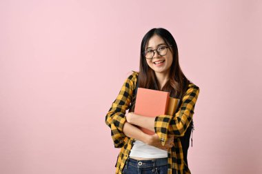 Güzel ve çekici Asyalı genç kız üniversite öğrencisi okul kitaplarını taşıyor ve soyutlanmış pembe bir arka plana karşı duruyor..