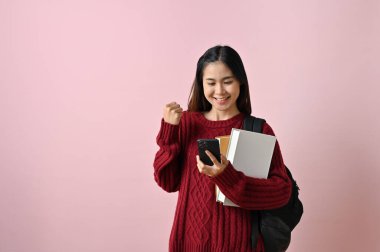 Akıllı telefon ekranına bakan genç Asyalı kız öğrenci yumruğunu sıkmış, jestini kutluyor, beklenmedik bir mesaj alıyor pembe izole bir arka plana karşı duruyor..