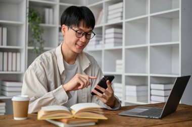 Genç yakışıklı Asyalı adam akıllı telefon kullanmayı seviyor, oturma odasında kulaklık takıyor.. 