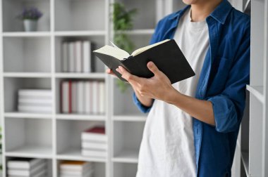 Çalışma odasında gözlüklü bir üniversite öğrencisinin kitap okuyup kitap rafına yaslanışını yakından izle.. 