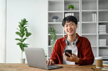 Çalışırken bilgisayar ve akıllı telefon kullanan neşeli genç bir üniversite öğrencisi..