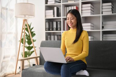 Genç, sevimli Asyalı kadın oturma odasında kanepede otururken dizüstü bilgisayar kullanıyor..