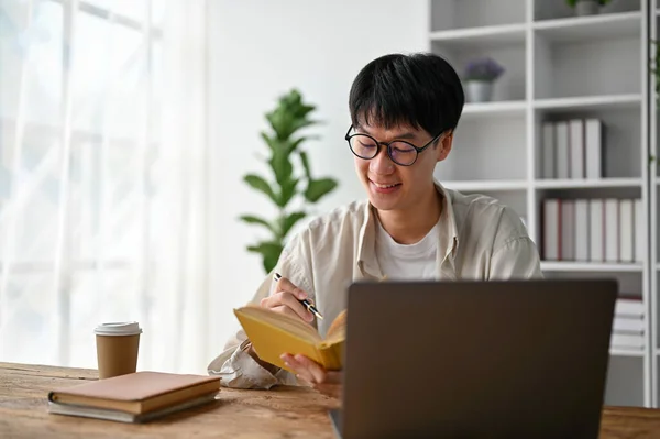 stock image Portrait of young Asian man writing on notebook while working with laptop computer in the living room.