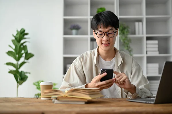 Stock image Handsome college student using smartphone in the living room, sitting at his study table. 