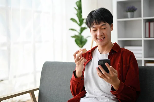 Schöner Junger Asiatischer Mann Isst Donut Und Benutzt Sein Smartphone — Stockfoto