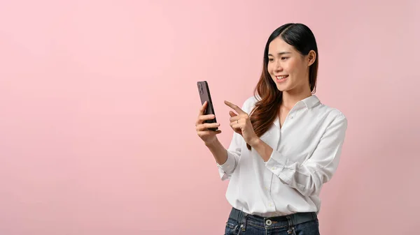 快乐的年轻亚洲女人指着被粉色背景隔离的智能手机 — 图库照片