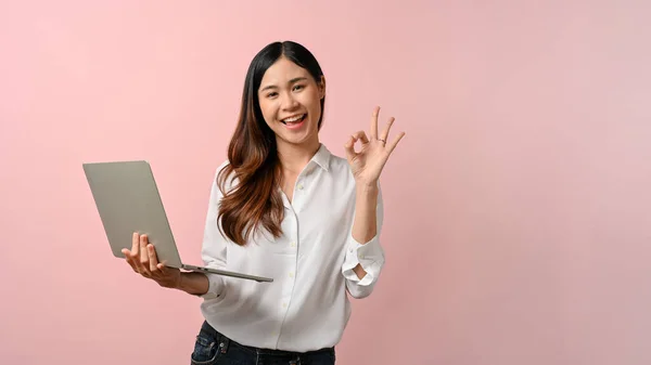 stock image Attractive and happy young Asian female in a white shirt with her laptop showing the okay hand sign, isolated on a pink background. Yes, agreement, perfect gesture