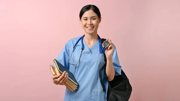 年轻美丽的亚洲医科学生拿着一个包和一本书对着摄像机微笑 与粉色背景隔离地站在一起 — 图库照片