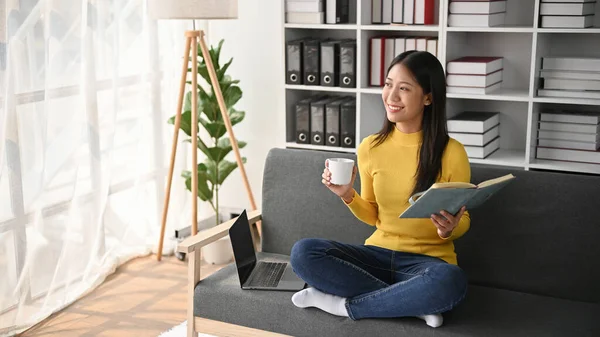 年轻的亚洲女人边喝咖啡边在客厅看书 享受闲暇的生活 — 图库照片