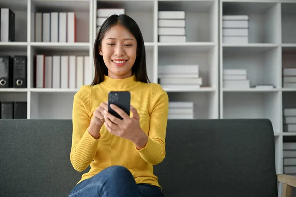快乐的年轻亚洲女人坐在沙发上 一边用智能手机 — 图库照片