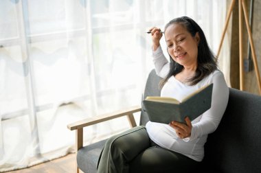 Güzel yaşlı bayan oturma odasındaki kanepede otururken boş zamanlarını kitap okuyarak geçiriyor.. 