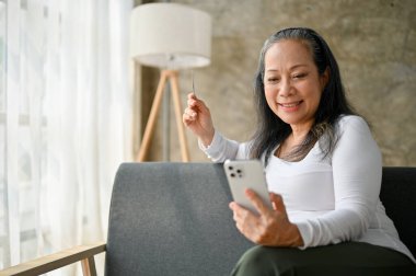 Kanepede otururken kredi kartıyla online alışveriş yapan mutlu yaşlı kadın..