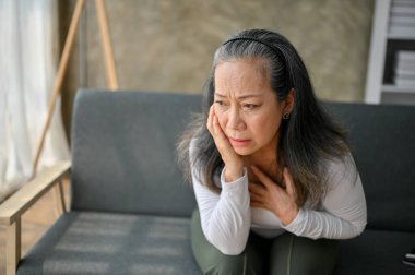 Şok olmuş ve endişeli Asyalı olgun kadın, oturma odasında koltukta otururken bir şey hakkında düşünüyor ve endişeleniyor, panik atak, endişe, depresyon.