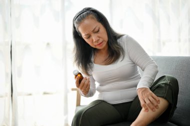 Mutsuz, olgun bir kadın bacak ağrısından muzdarip, evdeki kanepede bacağına masaj yapıyor. Sağlık sorunları kavramı
