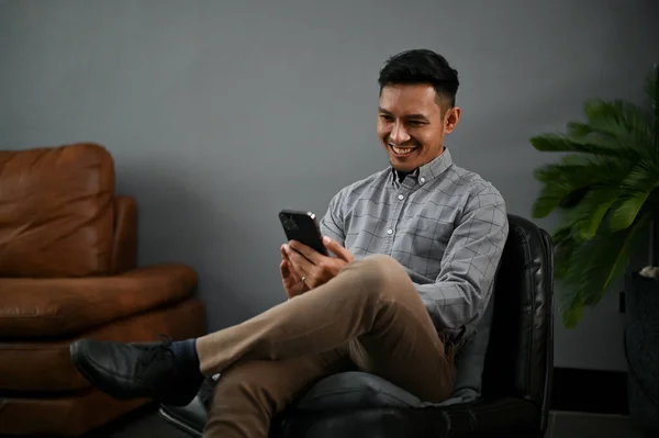 一个快乐的千年亚洲人在客厅里一边用智能手机 一边和别人聊天 一边在社交媒体上打滚 — 图库照片