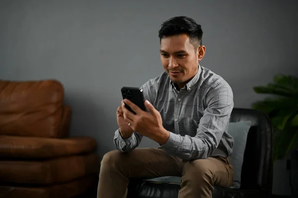一个英俊的男人坐在客厅里用他的智能手机查看他的电子邮件或短信 — 图库照片