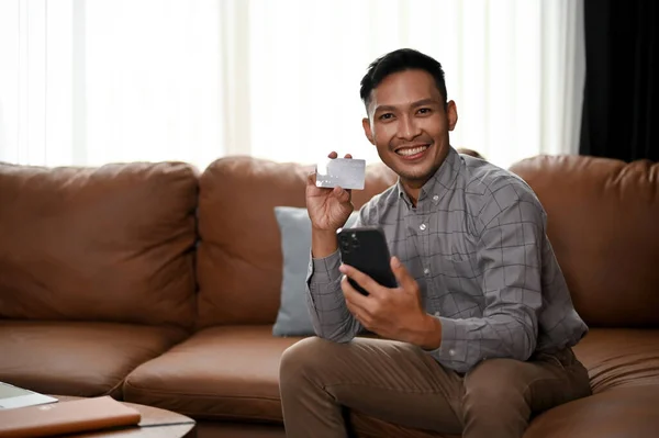 在客厅的沙发上 一个英俊 面带微笑的千年亚洲男人拿着智能手机 拿着信用卡拍照 — 图库照片