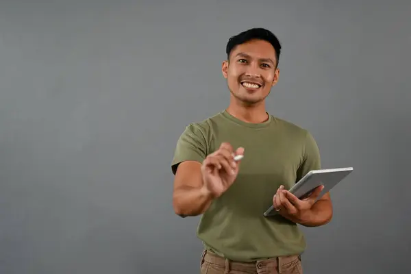 身穿休闲装 面带笑容的千年亚洲男子站在灰色的工作室背景上 手持数字平板电脑 用笔对着相机 — 图库照片