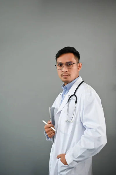 独立した灰色の背景に立って聴診器を持つ白いガウンでスマートでプロフェッショナルな千年のアジアの男性医師の肖像 — ストック写真
