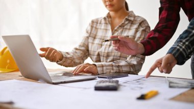 Ofiste mühendislerle birlikte çalışan, dizüstü bilgisayar ve plan üzerinde birlikte çalışan bir kadın mimarın kırpılmış görüntüsü..