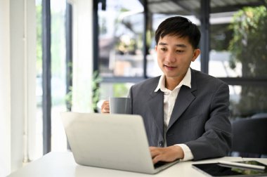 Zeki ve başarılı Asyalı iş adamı ya da erkek patron masasında dizüstü bilgisayarını kullanırken kahvesini yudumluyor..