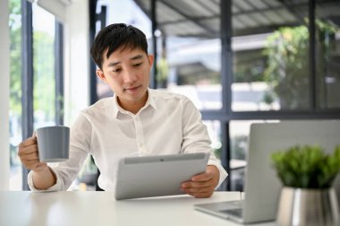 Yakışıklı ve zeki Asyalı iş adamı ya da beyaz gömlekli erkek patron tabletini kullanıyor ve modern bir ofiste masasında kahvesini yudumluyor..