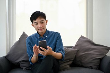 Yakışıklı ve mutlu bir Asyalı. Günlük kıyafetler içinde. Akıllı telefonunu oturma odasındaki bir kanepede kullanıyor..