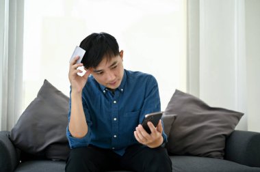 Milenyum Asyalı kafası karışmış bir adam oturma odasında bir koltukta oturuyor ve mobil bankacılık uygulamasında sorun yaşıyor..