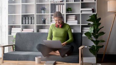 Mutlu ve gülümseyen, rahat giyimli Asyalı bir kadın, dizüstü bilgisayarını oturma odasındaki kanepede kullanarak arkadaşlarıyla sohbet etmekten hoşlanıyor..