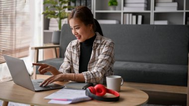 Çekici Asyalı kadın serbest yazar, modern oturma odasındaki bir kahve masasında dizüstü bilgisayardaki görevlerine odaklanıyor..