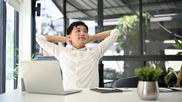 Relaxed millennial Asian businessman leaning on his office chair, stretching, and putting his arms behind his head to relax after finishing work.