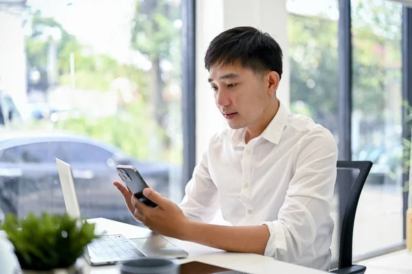 stock image Handsome and thoughtful millennial Asian businessman using his smartphone while sitting at his desk in the office. sms, chat, message, social media