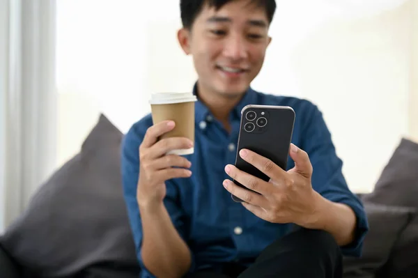 一个亚洲人在客厅里一边用智能手机一边喝着咖啡 选择性聚焦图像 — 图库照片