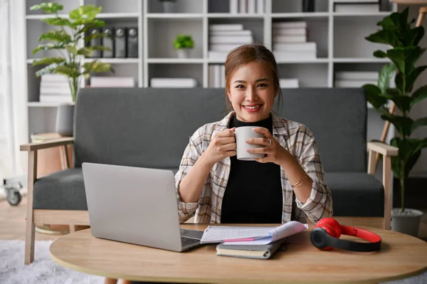 stock image Attractive and smiling young Asian female freelancer sits at a coffee table in her living room with a coffee cup, laptop and stuff. work from home concept