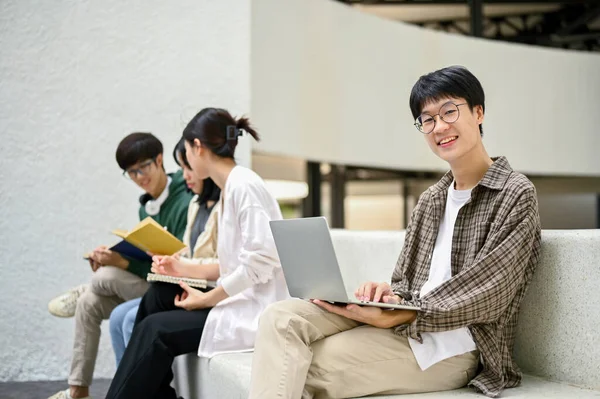 身穿休闲装的英俊而快乐的年轻亚洲大学生 拿着笔记本电脑坐在校园休闲区的长椅上 — 图库照片