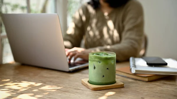 stock image Close-up image of a glass of iced matcha green tea on a wooden table in a minimal cafe with a female sitting behind.