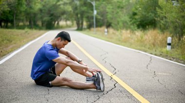 Atletik Asyalı bir adam koşarken kaslarına kramp giriyor, parkta bacağını esnetiyor. Yaralı, bacak ağrısı.