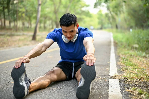 Handsome, athlete fit millennial Asian man stretching his legs and feet on the street, warming up before running at a nature park. Sport and leisure concept