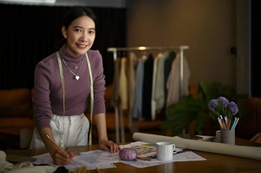 Gülümseyen ve büyüleyici Asyalı kadın moda tasarımcısı stüdyosunda, masasında duruyor..