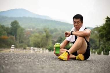 Yorgun ve susamış, spor giysili Asyalı bir adam halka açık bir parkta yolda oturuyor ve koşudan sonra kalp atışlarını ve kilometrelerce koşmasını kontrol ediyor..