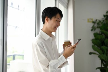 Beyaz gömlekli yakışıklı ve mutlu Asyalı genç ofis çalışanı akıllı telefonunu kullanarak pencerenin önünde duruyor, arkadaşlarıyla sohbet ediyor ve öğle yemeği arasında kahve içiyor..