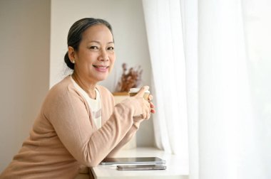 Gülümseyen ve çekici son sınıf Asyalı kadın rahat giysiler içinde kahve fincanını tutuyor, en az parlak kahve dükkanında oturuyor..