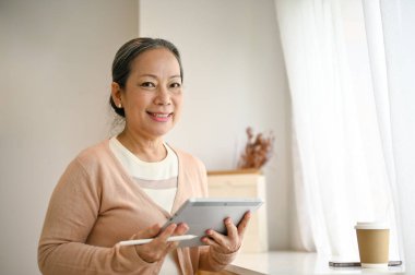 Gülümseyen ve çekici 60 yaşında sıradan kıyafetler giyen Asyalı bir kadın dijital tabletiyle küçük bir kafede oturuyor..