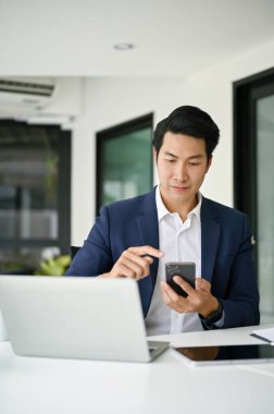 Çekici ve zeki Asyalı bir iş adamının portresi ya da profesyonel takım elbiseli bir erkek CEO 'nun akıllı telefonunu masasındaki tedarikçiye ulaşmak için kullanması..