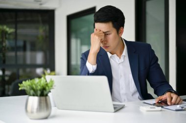 Stresli Asyalı iş adamı resmi takım elbisesiyle masasında oturuyor, sanata bakıyor dizüstü bilgisayarı ve sorunu çözmek için düşünceli bir plan yapıyor..