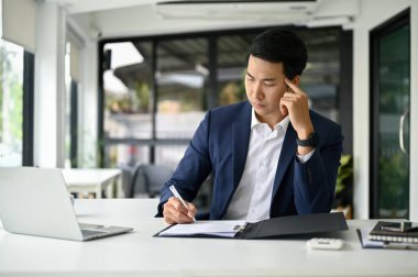 Profesyonel takım elbiseli Asyalı düşünceli bir işadamı mali raporları inceliyor ya da iş belgelerini tekrar kontrol ediyor, masasında çalışıyor..