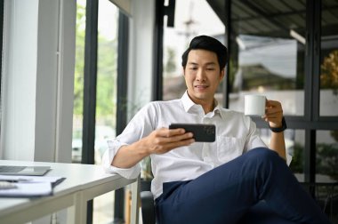 Gülümseyen ve rahat Asyalı iş adamı akıllı telefonuyla videolar izliyor ve sabah kahvesini ofisinde yudumluyor..