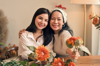 Güzel, çekici, orta yaşlı bir Asyalı anne ve güzel bir kızın portresi. Gülümseyen, sarılan ve güzel çiçeklerle dolu bir masada oturan..
